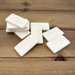 Puede incluir: Un juego de ocho fichas de domin&oacute; blancas hechas de hueso. Las fichas de domin&oacute; est&aacute;n dispuestas en una pila y esparcidas sobre una superficie de madera.