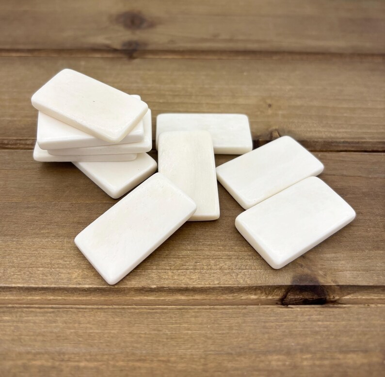 Puede incluir: Un juego de ocho fichas de domin&oacute; blancas hechas de hueso. Las fichas de domin&oacute; est&aacute;n dispuestas en una pila y esparcidas sobre una superficie de madera.