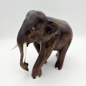 May include: A dark brown wooden elephant figurine with a polished finish. The sculpture features two smooth, light-colored tusks, carved facial details, and white accents on the feet. The elephant is depicted in a standing position.