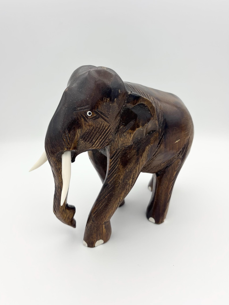 May include: A dark brown wooden elephant figurine with a polished finish. The sculpture features two smooth, light-colored tusks, carved facial details, and white accents on the feet. The elephant is depicted in a standing position.