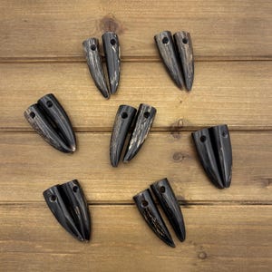 Peut inclure: Sept paires d'objets en forme de corne, brun foncé, avec un trou en haut. Les objets ont une finition polie et brillante et un motif de grain de bois naturel. Ils sont disposés sur une surface en bois.