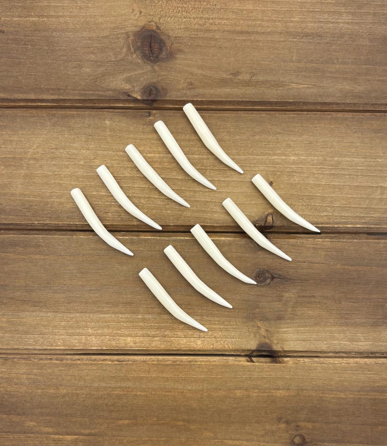 May include: A collection of nine smooth, off-white, curved, tapered horn-shaped components arranged in a diagonal pattern. Each piece features a rounded base and a pointed tip, presenting a uniform, solid, and polished appearance throughout the set.