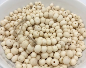 May include: A white ceramic bowl filled with cream-colored wooden beads of varying sizes. A strand of beads is strung on a natural twine. The beads have a matte finish and are scattered throughout the bowl.