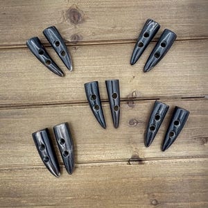 Może przedstawiać: Osiem czarnych guzików z rogu z dwoma otworami każdy. Guziki są ułożone w wzór na drewnianej powierzchni.