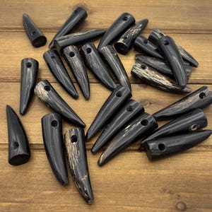 May include: A collection of dark brown, horn-shaped toggles with a hole at the top for sewing. Some toggles have a lighter, wood-grain pattern. The toggles are scattered on a wooden surface.