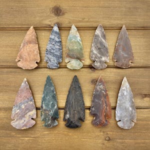 May include: Ten stone arrowheads arranged in two rows of five. Each arrowhead features a unique color and pattern, including shades of tan, grey, blue, green, black, and brown. The arrowheads have pointed tips and notched bases for attachment.