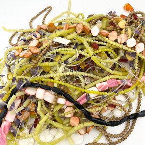 May include: A pile of various beaded strands in shades of green, brown, black, pink, and orange. The beads are of different shapes and sizes, including round, rectangular, and flat disc shapes. Some strands are knotted, and others are loose.
