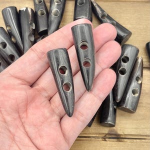 Könnte beinhalten: Nahaufnahme einer Hand, die eine Gruppe schwarzer Hornknöpfe mit jeweils zwei Löchern hält. Die Knöpfe sind spitz oval geformt und haben eine glatte, polierte Oberfläche.