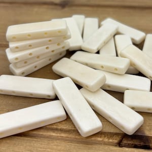 May include: Cream-coloured rectangular beads with rounded edges and three holes. Some beads are stacked, others scattered on a wooden surface. These beads are suitable for jewellery making and crafts.