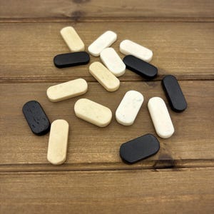 May include: A collection of small, rectangular wooden game pieces in various colors, including black, white, and cream. The pieces are scattered on a wooden surface, creating a visually appealing arrangement. The rounded edges of the pieces are visible.