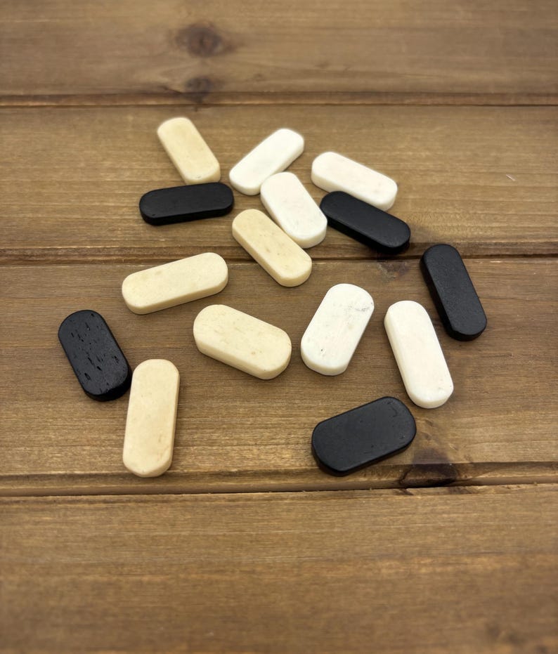 May include: A collection of small, rectangular wooden game pieces in various colors, including black, white, and cream. The pieces are scattered on a wooden surface, creating a visually appealing arrangement. The rounded edges of the pieces are visible.
