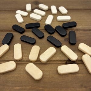 May include: A collection of small, rectangular wooden game pieces in three colors: black, off-white, and natural wood. The pieces are scattered on a brown wooden surface, suggesting a board game or craft project.