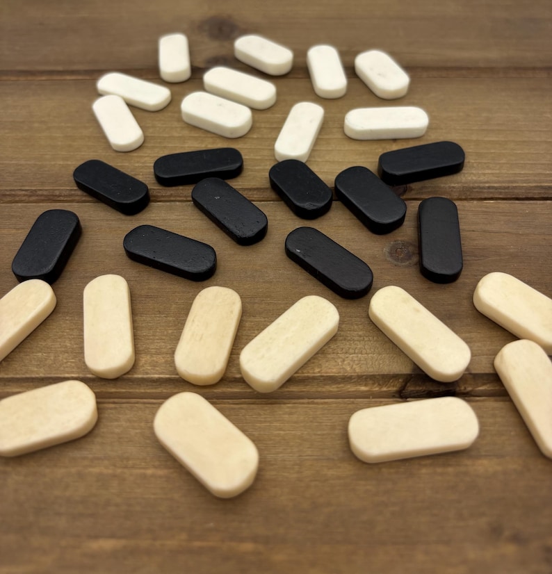 May include: A collection of small, rectangular wooden game pieces in three colors: black, off-white, and natural wood. The pieces are scattered on a brown wooden surface, suggesting a board game or craft project.