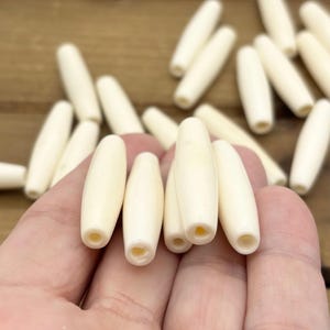 May include: A collection of off-white, elongated, cylindrical beads with a smooth finish and a central hole running through the length of each piece. The beads are tapered at both ends and have a consistent, solid, light cream color throughout.