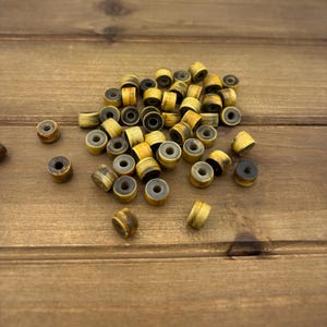 May include: A collection of small, cylindrical wooden beads with a light brown and dark brown striped pattern. Each bead has a gray center hole. The beads are scattered on a wooden surface.