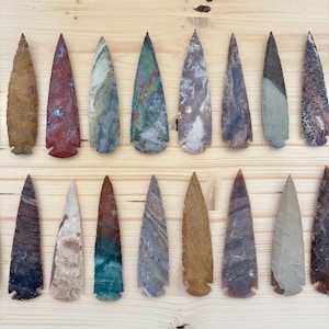 May include: A collection of sixteen stone arrowheads arranged in two rows. Each arrowhead features a unique color, including shades of red, blue, green, brown, grey, and tan, with varied textures and patterns across the pointed stone surfaces.