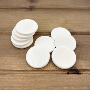 Puede incluir: Una pila de diez posavasos redondos de madera blanca. Los posavasos son lisos y tienen un acabado ligeramente brillante.