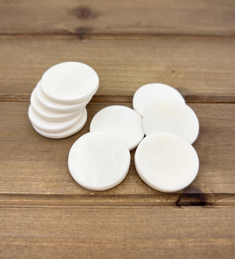 Puede incluir: Una pila de diez posavasos redondos de madera blanca. Los posavasos son lisos y tienen un acabado ligeramente brillante.