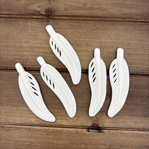 May include: Five white ceramic feathers with a cut-out design. The feathers are arranged in a semi-circle on a wooden surface.