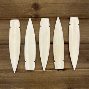 May include: Five white bone carving tools with pointed tips and a rounded edge. The tools are arranged in a row on a wooden surface.