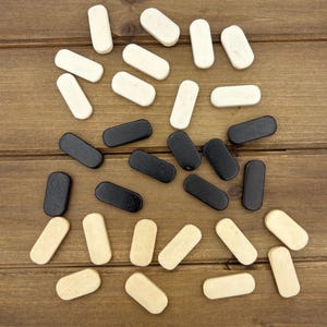 May include: Overhead shot of wooden game pieces in three colors: black, off-white, and natural wood. The pieces are rectangular with rounded ends, scattered on a brown wooden surface. The game pieces are likely for a board game.