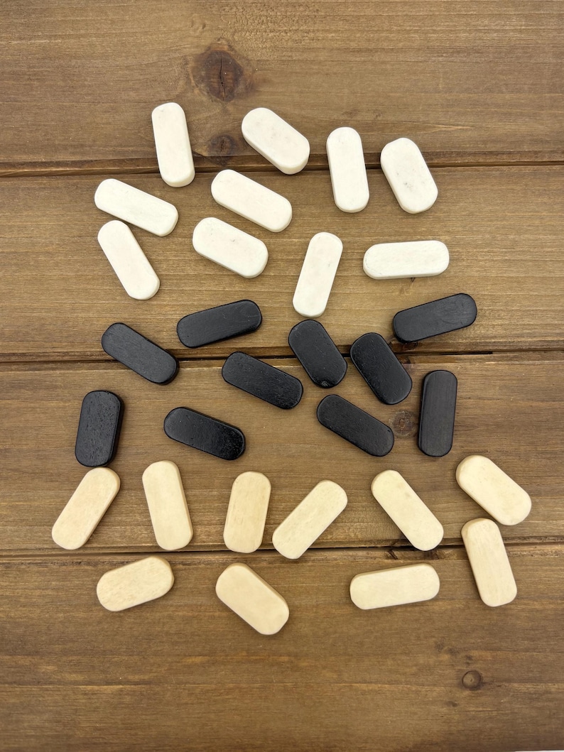 May include: Overhead shot of wooden game pieces in three colors: black, off-white, and natural wood. The pieces are rectangular with rounded ends, scattered on a brown wooden surface. The game pieces are likely for a board game.