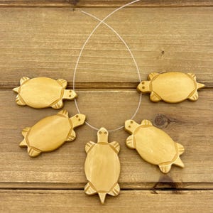 May include: A necklace featuring five wooden turtle pendants. The turtles are light brown with carved details and are strung on a thin, clear cord. The background is a wooden surface.