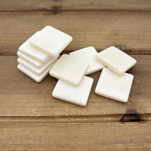 Puede incluir: Un mont&oacute;n de seis azulejos cuadrados blancos sobre una superficie de madera. Los azulejos son lisos y tienen un acabado ligeramente brillante.