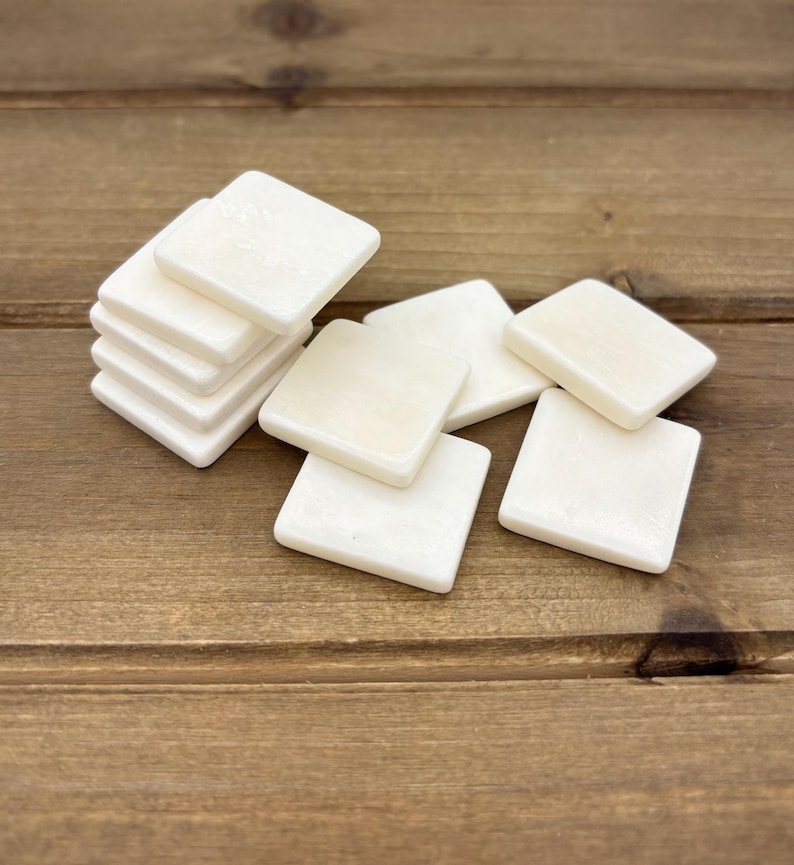 Puede incluir: Un mont&oacute;n de seis azulejos cuadrados blancos sobre una superficie de madera. Los azulejos son lisos y tienen un acabado ligeramente brillante.