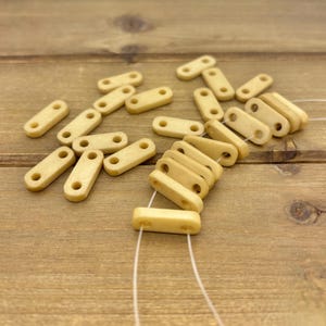 Könnte beinhalten: Kleine, rechteckige, cremefarbene Perlen mit zwei Löchern, auf einer Holzoberfläche verstreut. Einige sind mit weißem Faden aufgefädelt. Diese kleinen Perlen sind wahrscheinlich für die Schmuckherstellung oder Bastelarbeiten gedacht. Die Perlen haben eine glatte, matte Oberfläche.