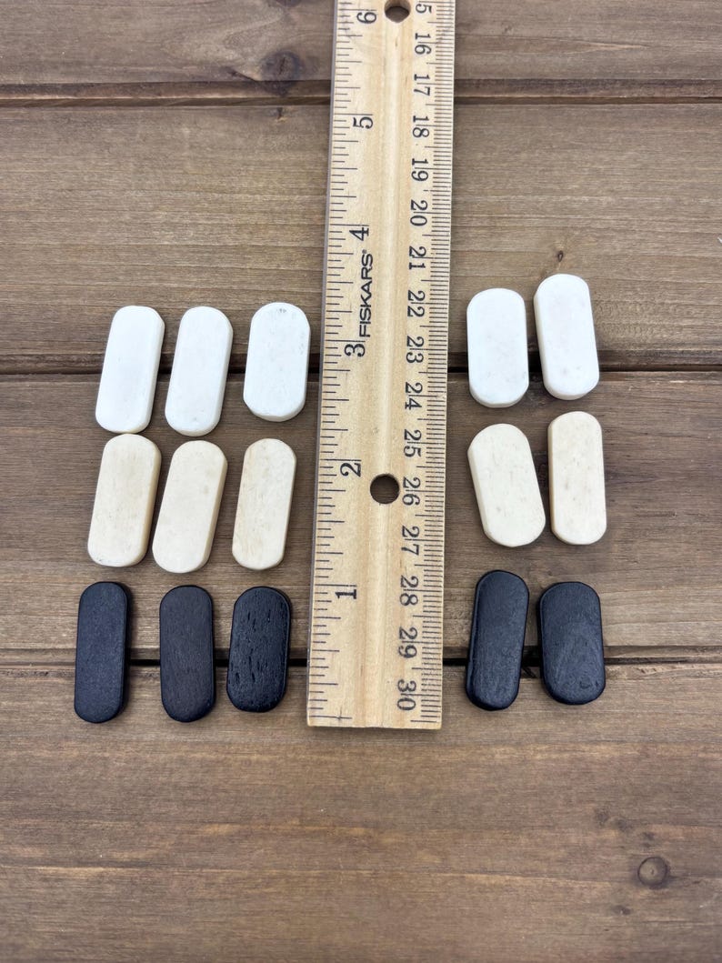 May include: A collection of small, rectangular wooden pieces in white, natural, and black arranged on a wooden surface. A ruler is placed vertically next to the pieces, showing measurements in inches.