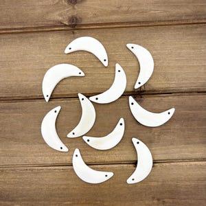 May include: A collection of white ceramic crescent moon shapes with small holes on the sides. The moon shapes are arranged in a circular pattern on a brown wooden surface.