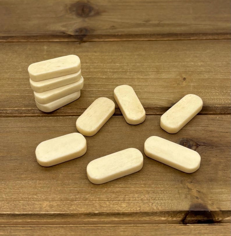 May include: A collection of small, rectangular, off-white game pieces. Some are stacked, while others are scattered on a wooden surface. The pieces have rounded edges and a smooth texture, suggesting they are made of bone or a similar material.