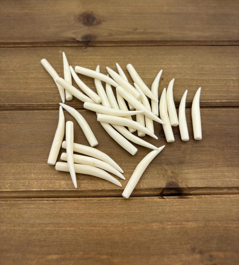 May include: A collection of numerous small, smooth, off-white, curved, tapered, tooth-shaped components. Each piece features a rounded base that narrows to a sharp, pointed tip, arranged in a scattered pile with varying orientations.