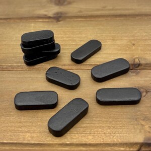 May include: A collection of small, black, rectangular wooden pieces with rounded edges. Some pieces are stacked, while others are scattered on a wooden surface. The pieces have a smooth, matte finish and are uniformly colored.