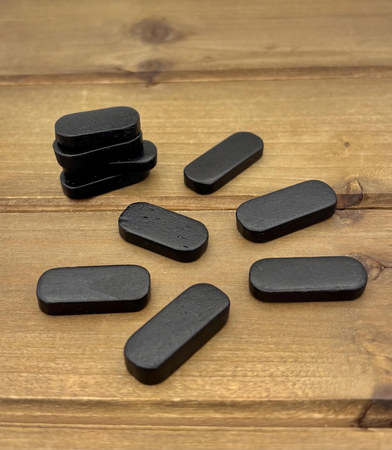 May include: A collection of small, black, rectangular wooden pieces with rounded edges. Some pieces are stacked, while others are scattered on a wooden surface. The pieces have a smooth, matte finish and are uniformly colored.