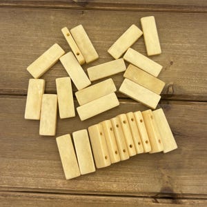 May include: A collection of small, rectangular, off-white wooden beads and blocks. Some blocks have holes drilled through them. The items are scattered on a brown wooden surface.