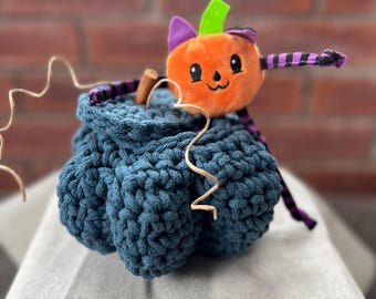 May include: A crocheted pumpkin in a teal blue color with a small plush pumpkin with a purple hat and green stem on top. The plush pumpkin has a cute face with eyes, a nose, and a smile.