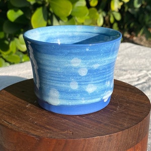 Puede incluir: Una taza pequeña de cerámica azul con motas blancas y un esmalte azul claro. La taza está sobre un soporte de madera.