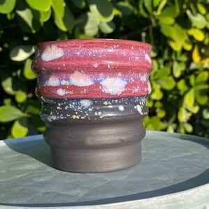 Puede incluir: Una taza pequeña de cerámica negra con un esmalte rojo y blanco. La taza tiene un diseño ondulado y texturizado y está sentada sobre una superficie metálica.