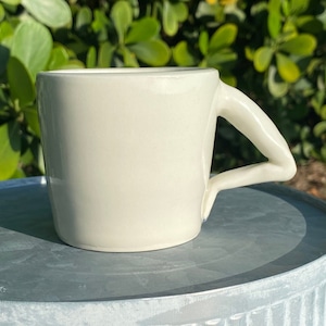 Puede incluir: Una taza de cerámica blanca con un asa curva única. La taza está sobre una superficie de metal gris.