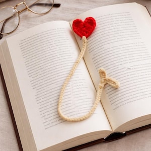 May include: An open book with a red crocheted heart bookmark. The bookmark is attached to a cream-colored cord. Gold-rimmed glasses rest on the page. A small potted plant is in the background. The book has a brown cover.