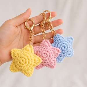 Op de afbeelding: Drie stervormige gehaakte sleutelhangers in pastelgeel, lichtroze en babyblauw. Elke ster is bevestigd aan een goudkleurige metalen karabijnhaak en een korte ketting, met een gestructureerd, cirkelvormig garensteekpatroon op de voorzijde.
