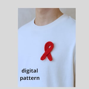 Puede incluir: Un broche de lazo rojo de ganchillo, un símbolo de concienciación, está prendido a una camiseta blanca. Las palabras "digital pattern" están impresas en negro debajo del lazo. La imagen está sobre un fondo gris claro.