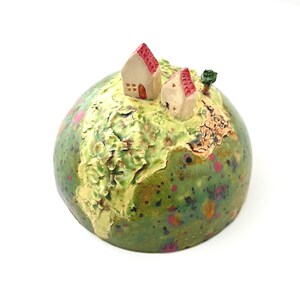 May include: A ceramic globe sculpture with two small houses and a tree on top. The globe is primarily green with textured, light green and tan areas. The houses have red roofs and tan walls. Pink and orange speckles dot the globe's surface.