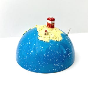 Op de afbeelding: Een keramische globe sculptuur met een blauw, gespikkeld oppervlak. Een gele landmassa met een rood-witte vuurtoren en een klein huisje staat bovenop de globe. Een klein, wit, puntig object is aan de zijkant van de globe bevestigd.