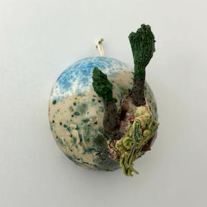 Op de afbeelding: Een bolvormig keramisch ornament met een geglazuurd oppervlak met blauwe, groene en crèmekleurige spikkels. Twee donkerbruine, getextureerde boomachtige structuren steken uit de bovenkant, met lichtgroene, wortelachtige ranken aan de zijkant.