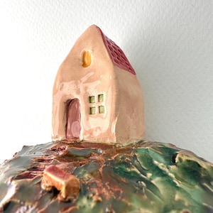 Może przedstawiać: Ceramiczna rzeźba przedstawiająca mały domek z jasnobrązową fasadą, czerwonym, teksturowanym dachem, ciemnoróżowymi drzwiami łukowymi i czterema małymi zielonymi oknami. Domek stoi na błyszczącej, nierównej podstawie w odcieniach zieleni, brązu i czerwieni.