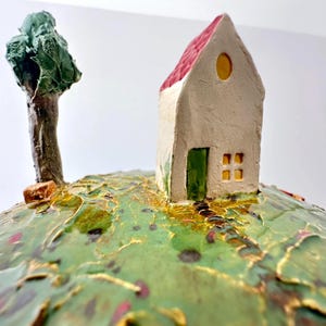 Op de afbeelding: Een keramisch beeldje van een klein wit huisje met een rood dak, een groene deur en gele ramen, geplaatst op een textuurrijke groene heuvel met gouden accenten. Een gebeeldhouwde groene boom staat links van het huis op het oppervlak.
