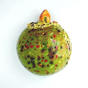 Puede incluir: Un adorno esférico de cerámica con una superficie verde brillante y motas rojas y marrón oscuro. Una pequeña casa naranja tridimensional con una puerta verde se asienta sobre la esfera, rodeada de detalles en relieve de color crema texturizados.